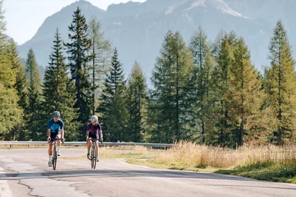 Two cyclists riding on a winding road through a sunny mountain forest