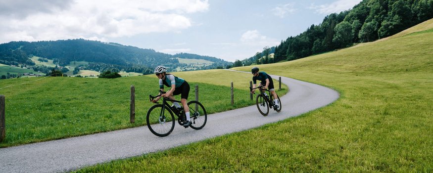 Zwei Radfahrer fahren auf einer kurvigen Landstraße durch grüne Hügel.