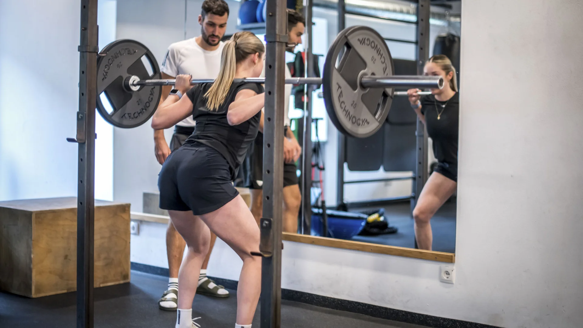 Frau macht Kniebeugen mit Langhantel im Fitnessstudio, betreut von Trainer