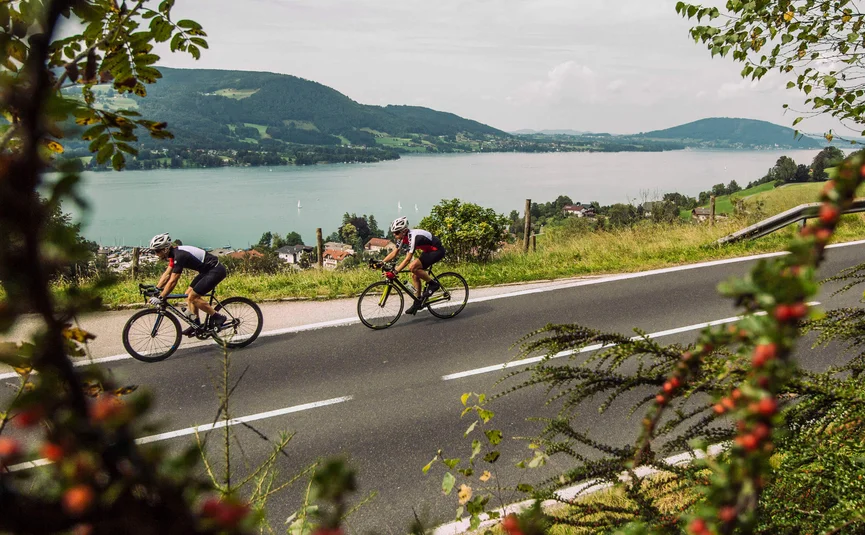 Zwei Radfahrer auf Landstraße mit Berg- und Seeblick