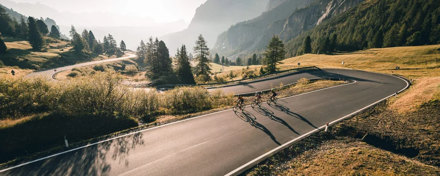 Alta Badia © Molography Tre ciclisti su strada di montagna curva al sole