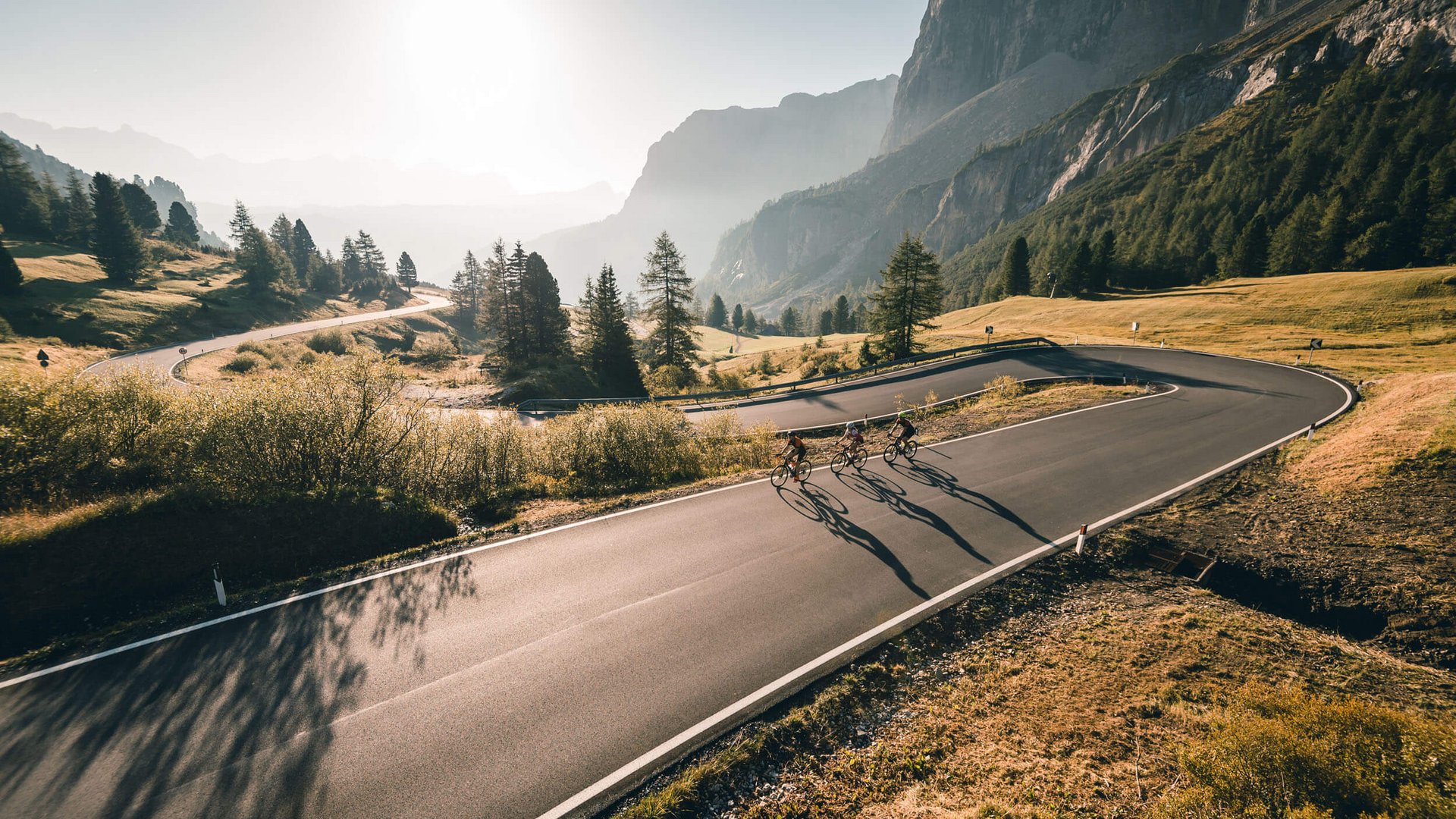 Three cyclists riding on a winding mountain road in sunlight