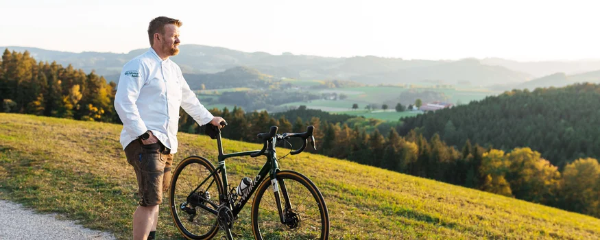 Mit Profis trainieren im Krumbacherhof © NOE Werbung - Mara Hohla Mann steht mit Rennrad am Weg und blickt auf hügelige Landschaft im Sonnenuntergang