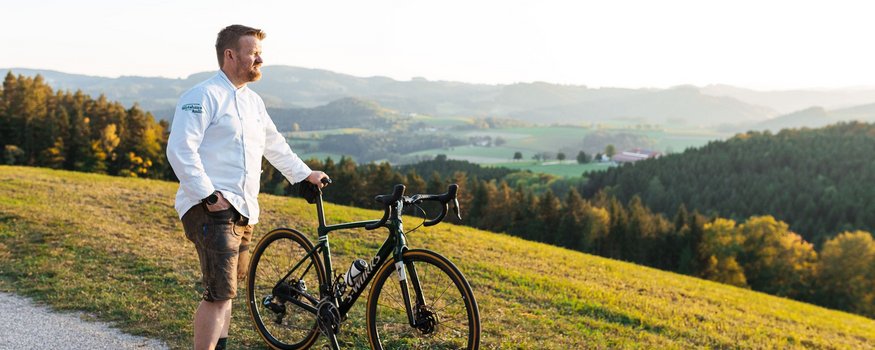 Mann steht mit Rennrad am Weg und blickt auf hügelige Landschaft im Sonnenuntergang
