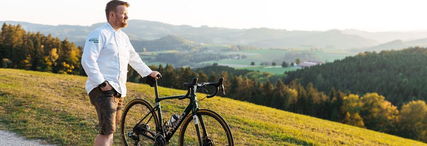 Mann steht mit Rennrad am Weg und blickt auf hügelige Landschaft im Sonnenuntergang