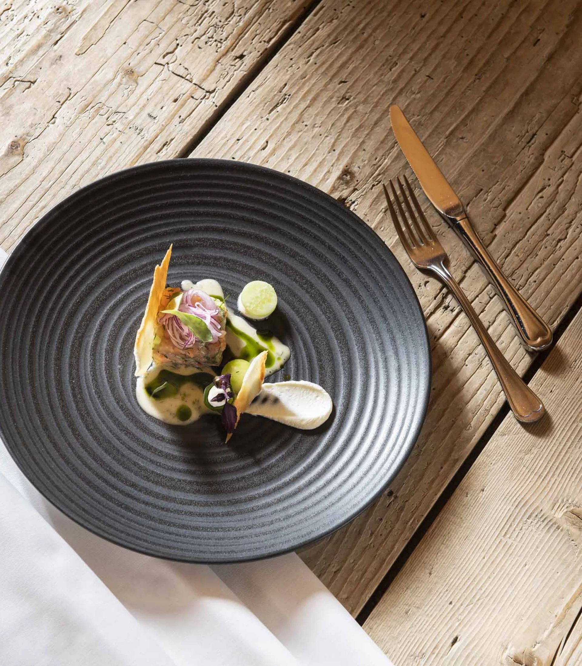 Piatto gourmet artisticamente impiattato su piatto nero con posate su tavolo di legno