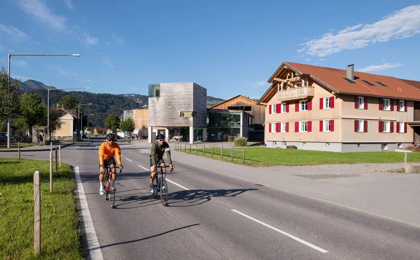 Twee wielrenners fietsen op een weg met gebouwen en bergen op de achtergrond