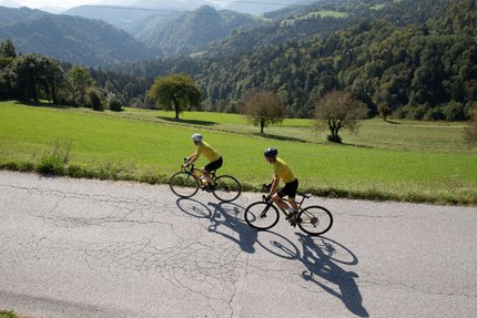 Due ciclisti su una strada tra colline verdi e montagne sullo sfondo