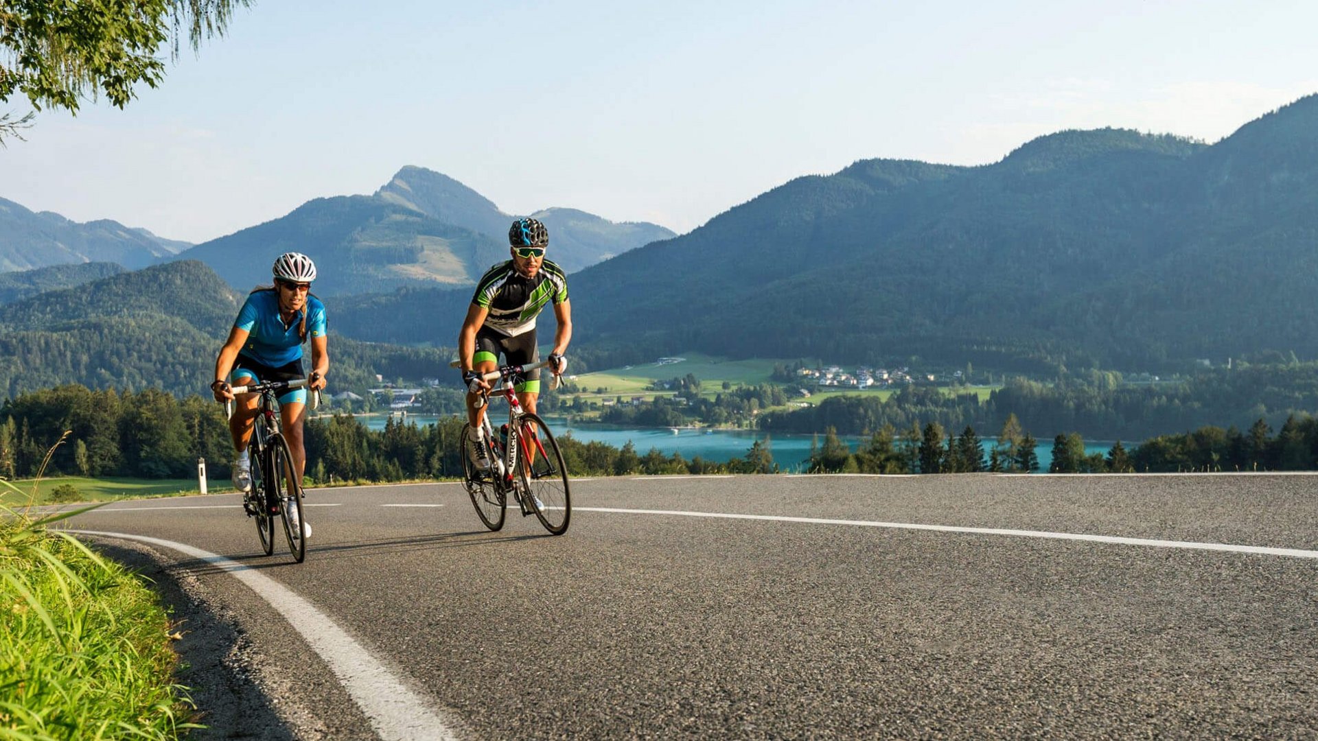 Zwei Radfahrer fahren bergauf auf einer Straße mit Bergen und See im Hintergrund