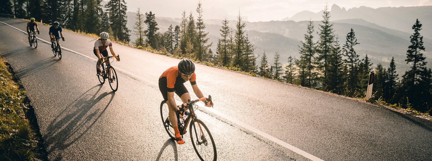 Four cyclists riding uphill on a road with trees and mountains in the background