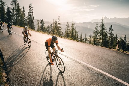 Four cyclists riding uphill on a road with trees and mountains in the background