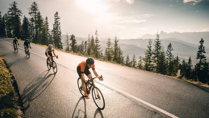 Vier Radfahrer fahren bergauf auf einer Straße mit Bäumen und Bergen im Hintergrund