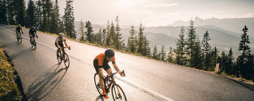 Four cyclists riding uphill on a road with trees and mountains in the background
