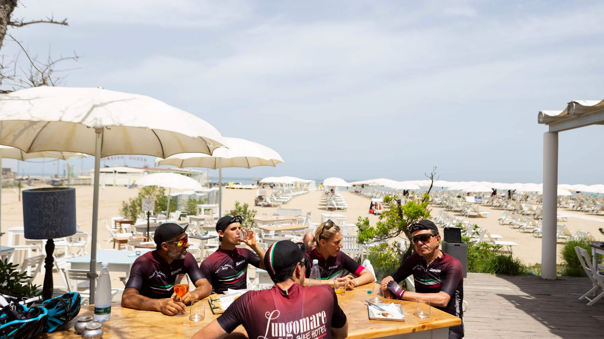 Cyclists in jerseys sitting at a wooden table under umbrellas on a beach