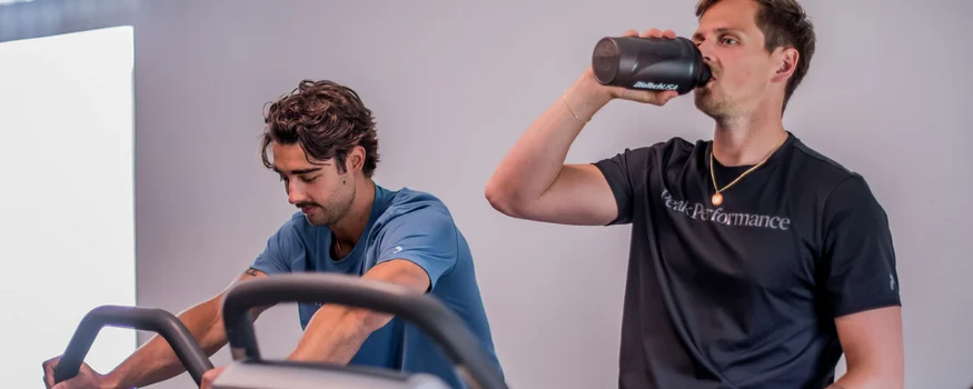 Mehr als Rennradurlaub: Sporthotel Zoll setzt auf Performance © Fabian Schaiter Zwei Männer trainieren auf Heimfahrrädern, einer trinkt aus einem Shaker