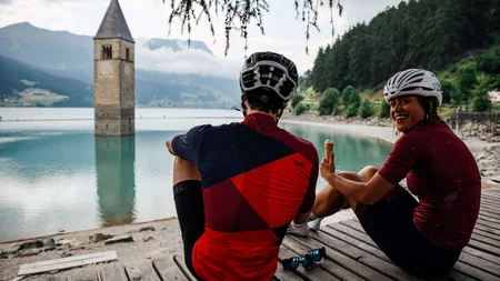 Zwei Radfahrer sitzen am See mit einem im Wasser stehenden Kirchturm
