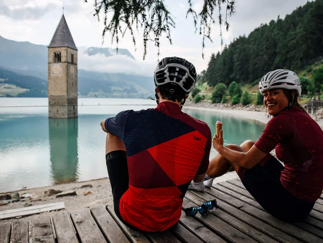 Zwei Radfahrer sitzen am See mit einem im Wasser stehenden Kirchturm