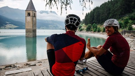 Zwei Radfahrer sitzen am See mit einem im Wasser stehenden Kirchturm