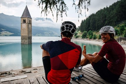 Due ciclisti seduti vicino a un lago con una torre della chiesa sommersa