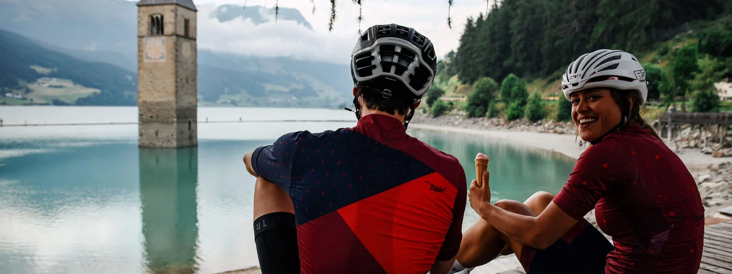 3 paesi Giro © Felix Saller Due ciclisti seduti vicino a un lago con una torre della chiesa sommersa