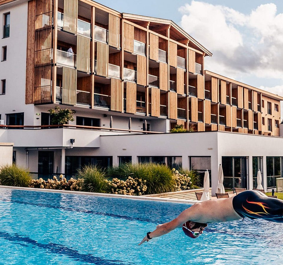 Bike.Move.Relax. im Das Hohe Salve Sportresort © Daniel Waschnig Mann taucht in einen Außenpool vor einem modernen Hotelgebäude ein