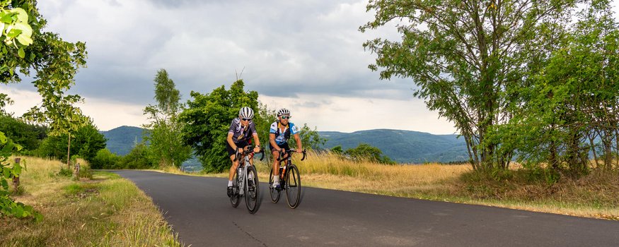 Erzgebirge © MountainloversTVE Zwei Radfahrer fahren auf einer ruhigen Landstraße mit Hügeln im Hintergrund