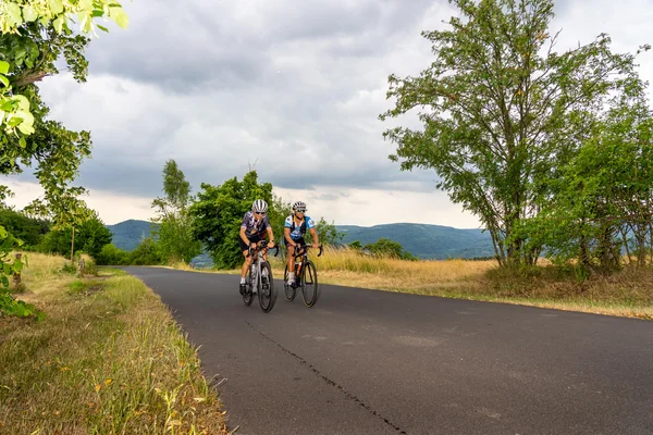 Deutschland © MountainloversTVE Zwei Radfahrer fahren auf einer ruhigen Landstraße mit Hügeln im Hintergrund