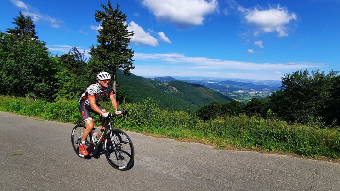 Ciclista che pedala in salita su strada panoramica con colline verdi e cielo blu