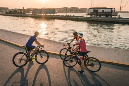 Three cyclists stopped by water at sunset with buildings in background