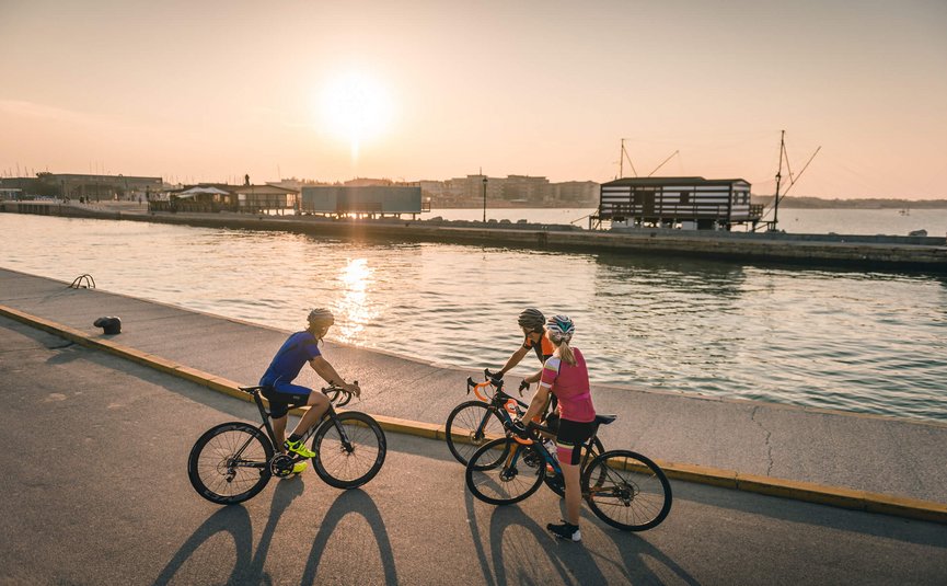 Three cyclists stopped by water at sunset with buildings in background