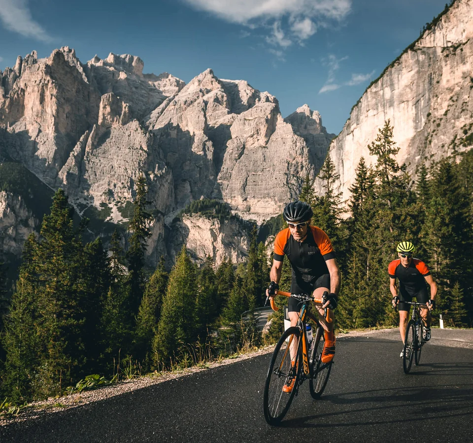 Warum Alta Badia auf jede Roadbike-Bucket-List gehört © Molography Zwei Radfahrer fahren auf Bergstraße mit Felsen und Nadelbäumen im Hintergrund