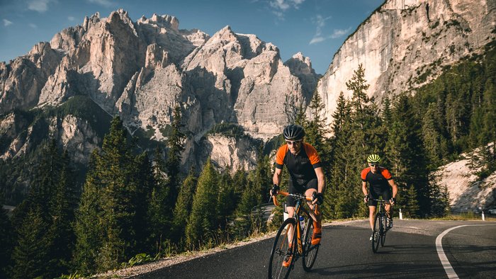 Zwei Radfahrer fahren auf Bergstraße mit Felsen und Nadelbäumen im Hintergrund