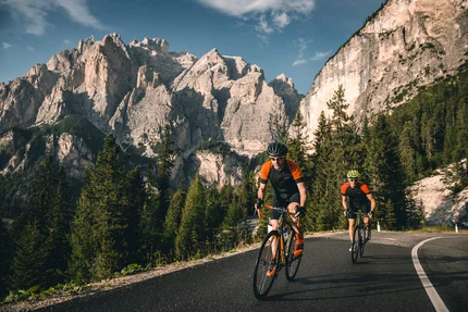 Warum Alta Badia auf jede Roadbike-Bucket-List gehört © Molography Zwei Radfahrer fahren auf Bergstraße mit Felsen und Nadelbäumen im Hintergrund