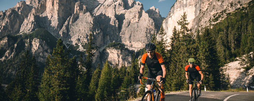 Two cyclists riding on a mountain road with rocky cliffs and pine trees behind