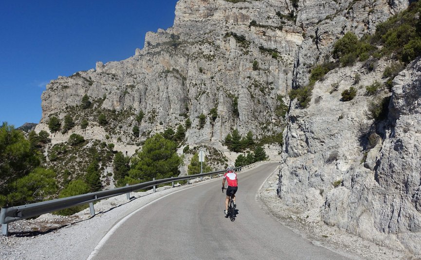 Ciclista su strada di montagna tortuosa con scogliere rocciose e cielo blu