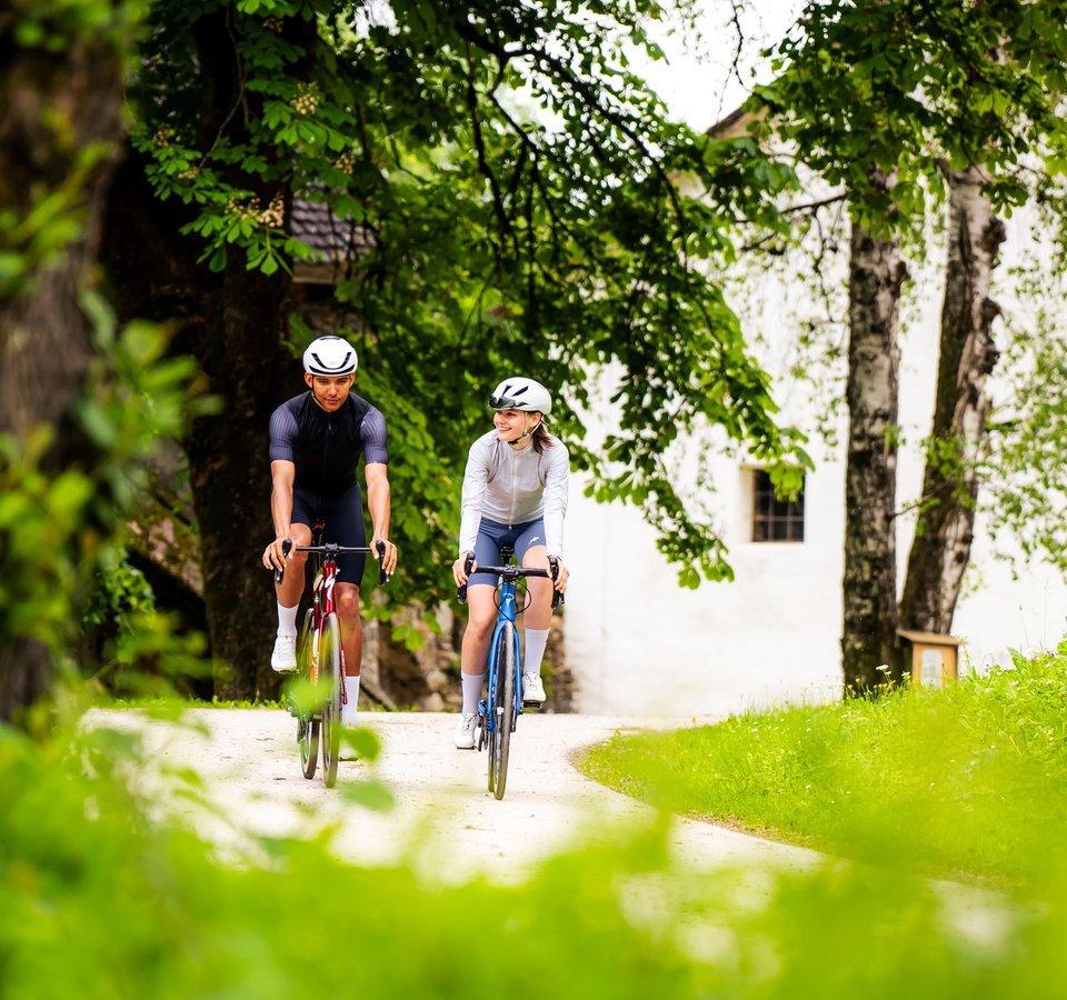 Zwei Fahrradfahrer auf einem Waldweg neben Bäumen und einer weißen Wand