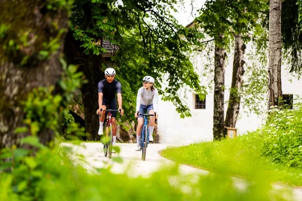 Zwei Fahrradfahrer auf einem Waldweg neben Bäumen und einer weißen Wand