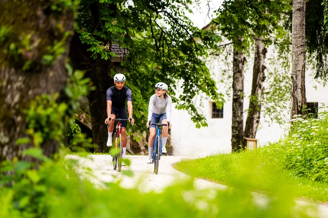 Zwei Fahrradfahrer auf einem Waldweg neben Bäumen und einer weißen Wand