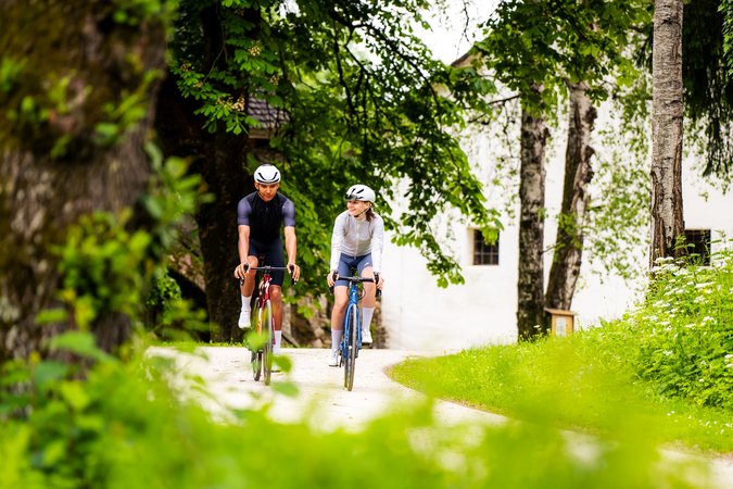 Zwei Fahrradfahrer auf einem Waldweg neben Bäumen und einer weißen Wand