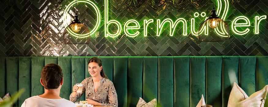 Paar sitzt im gemütlichen Café mit grünem Sofa und Obermüller Leuchtschrift