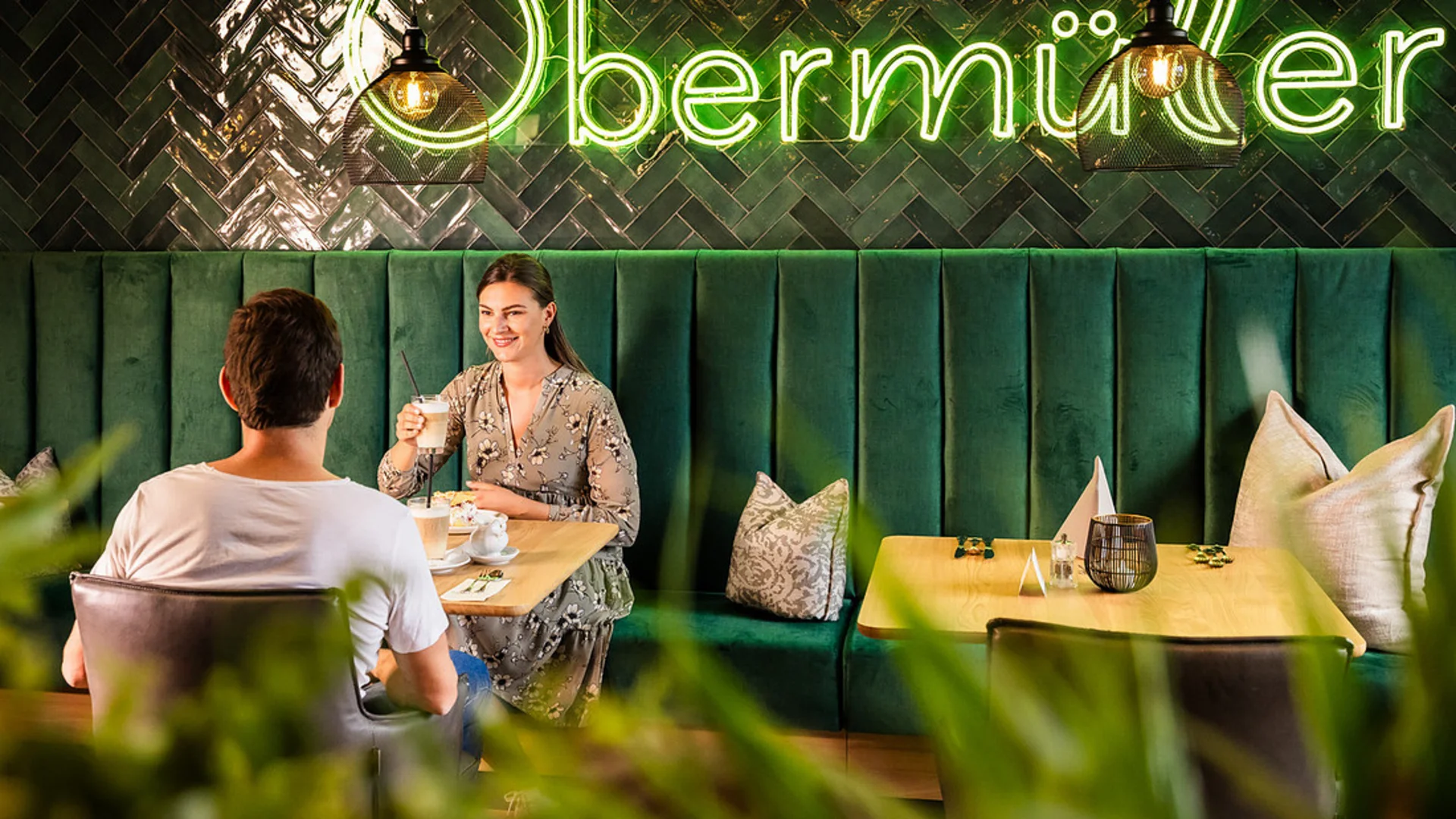Couple seated in cozy café with green sofa and Obermüller neon sign