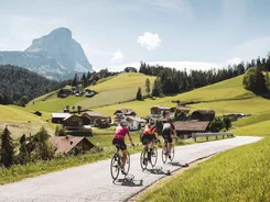 Three cyclists ride on rural road in sunny valley with mountain backdrop