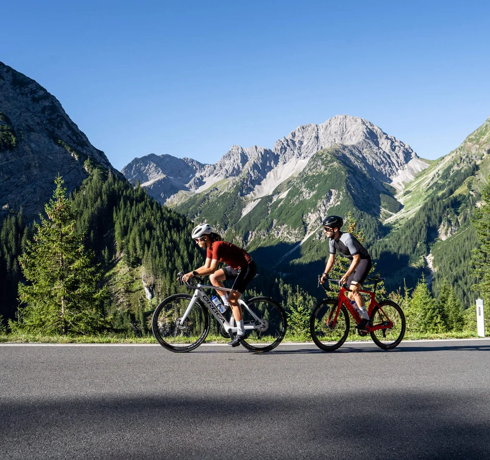 Zwei Radfahrer fahren auf Bergstraße mit Alpen im Hintergrund