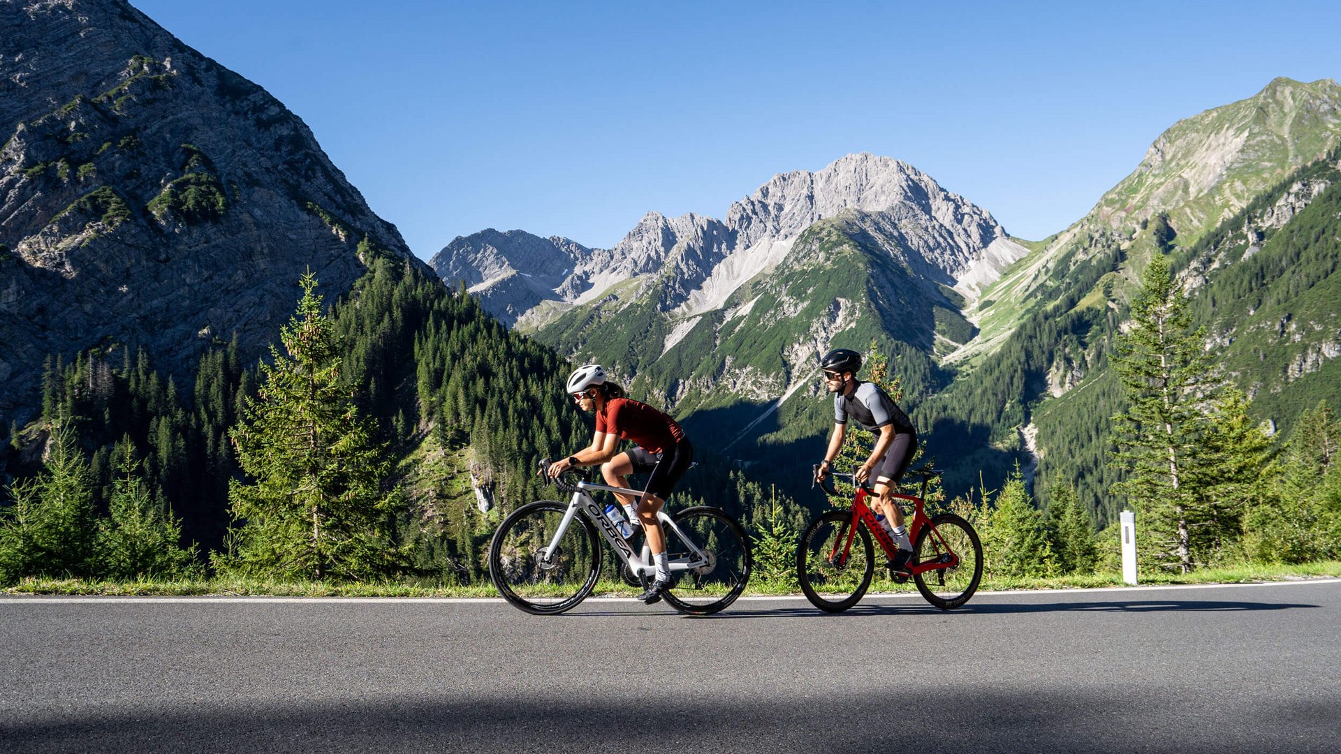 Zwei Radfahrer fahren auf Bergstraße mit Alpen im Hintergrund