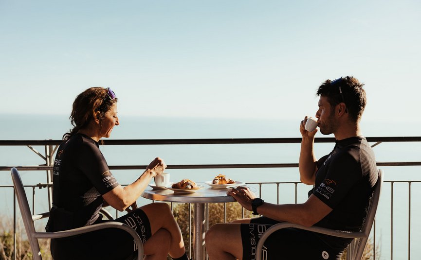 Due ciclisti che bevono caffè e mangiano cornetti su balcone con vista mare