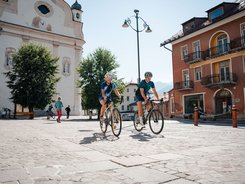 Due ciclisti pedalano su una piazza con edifici storici in una giornata di sole