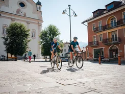 Due ciclisti pedalano su una piazza con edifici storici in una giornata di sole