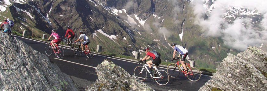 Cyclists riding uphill on mountain road with snowy Alps in background