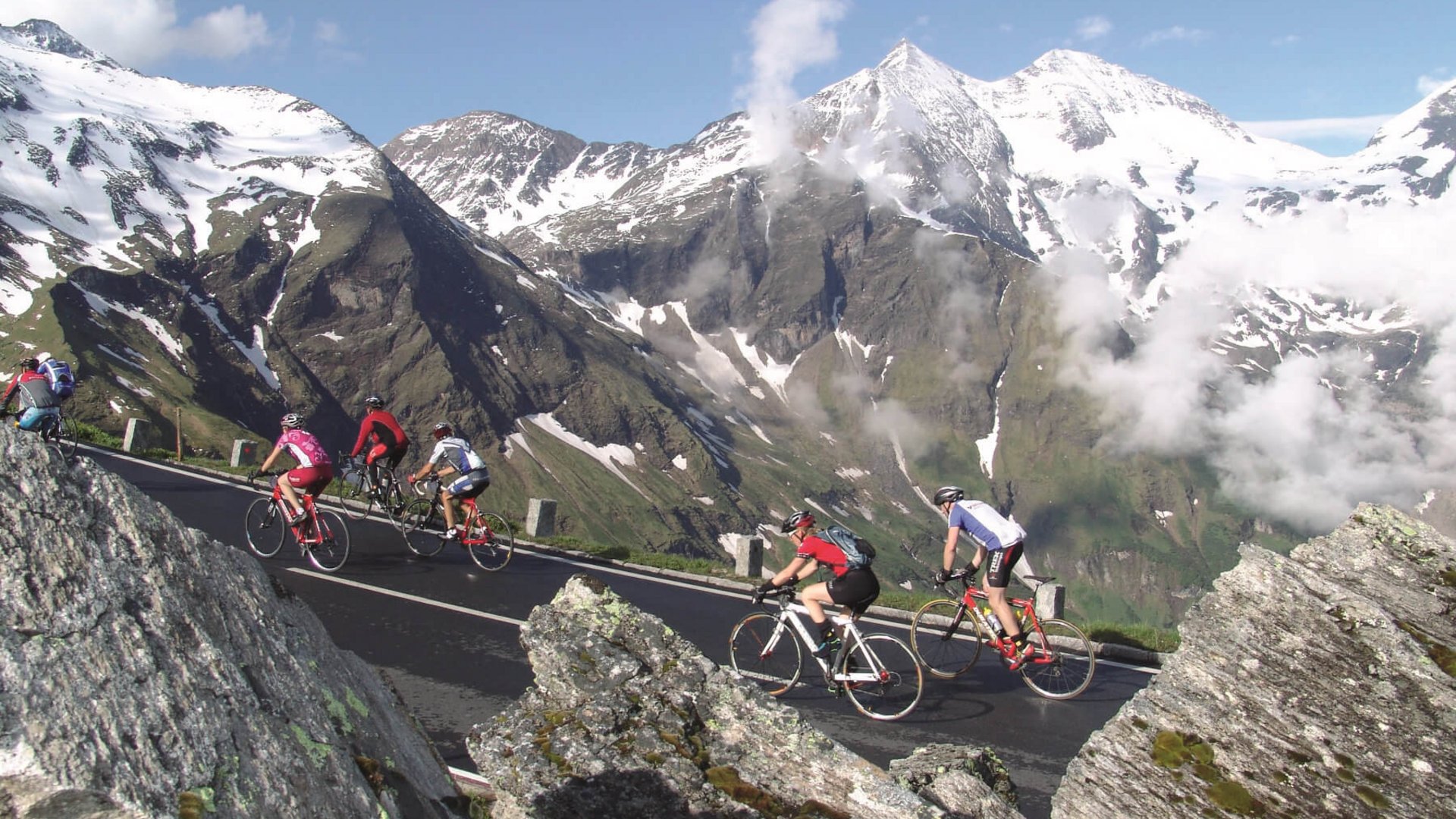 Triathlon Hotels: Deine Base für Training & Erholung © Sportcamp Woferlgut Bruck Radfahrer fahren auf Bergstraße mit schneebedeckten Alpen im Hintergrund
