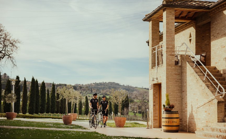 Due ciclisti in abbigliamento nero camminano con le loro bici accanto a un edificio in mattoni